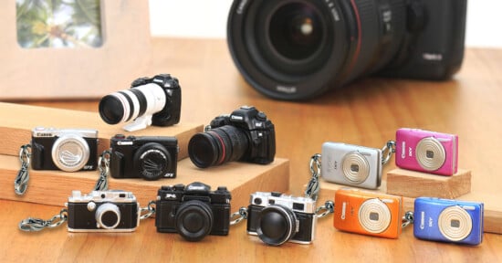 A collection of miniature Canon camera keychains arranged on a wooden surface in front of a full-sized camera lens.