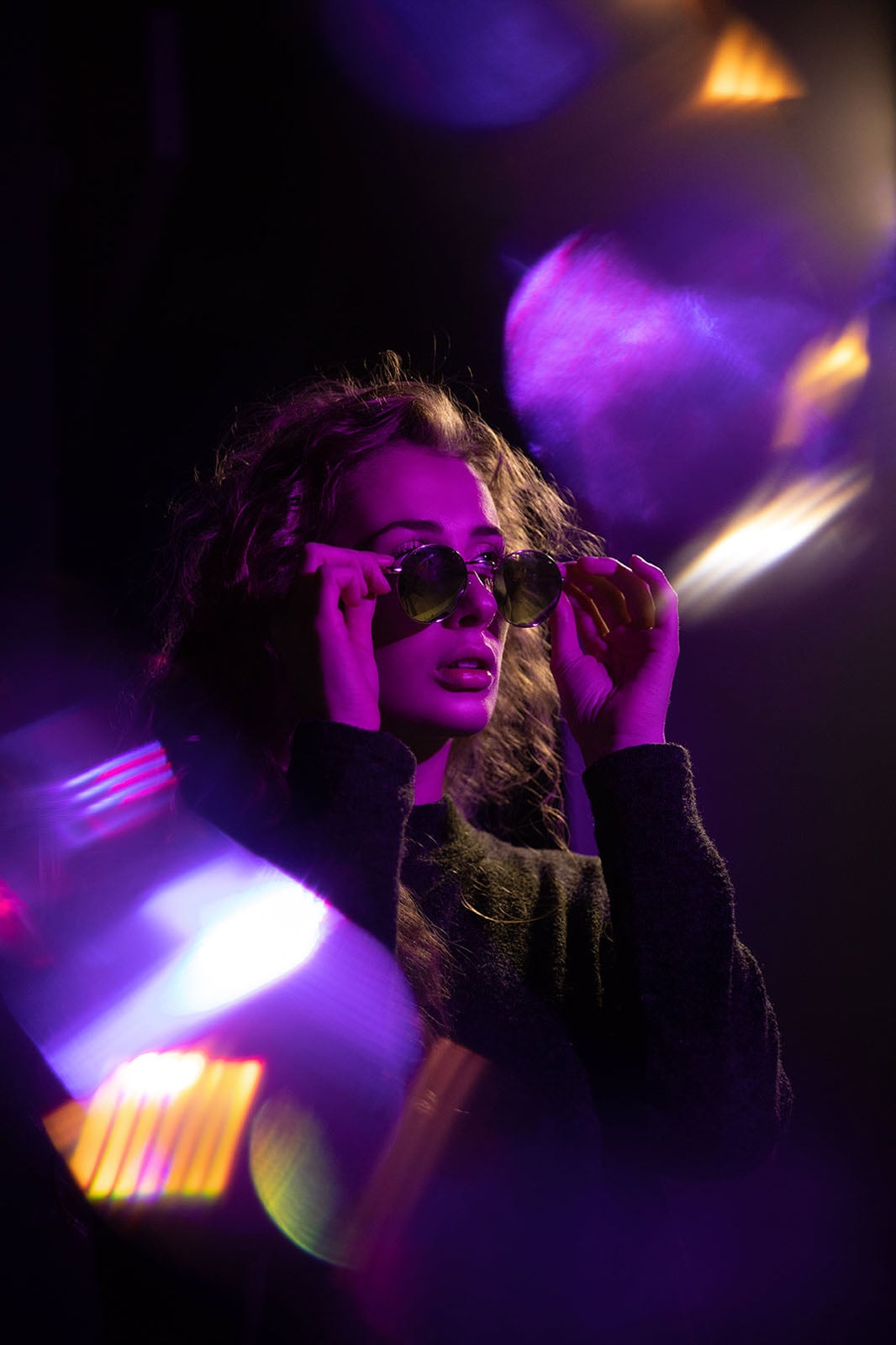 A woman with curly hair wears round sunglasses while posing in a dark environment with vibrant purple and orange light flares.