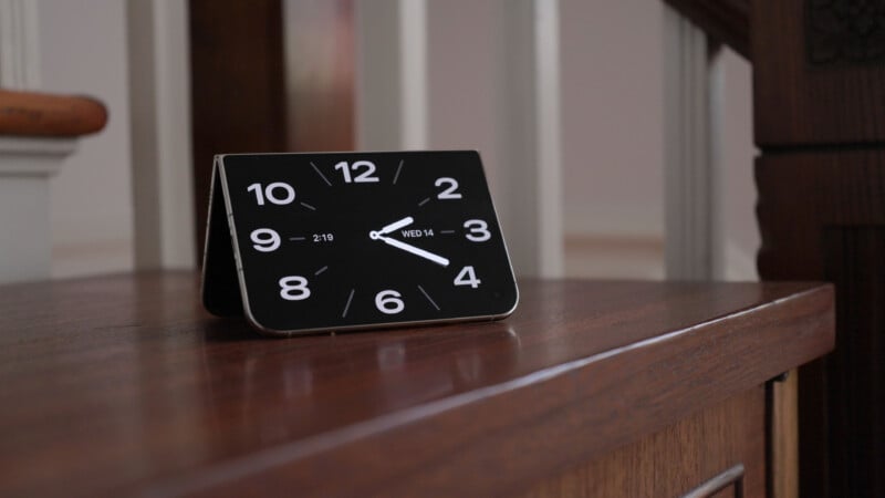 A foldable smartphone sits on a wooden surface in a tented position, displaying a digital clock face with the time 4:20 and the date Wednesday the 14th.