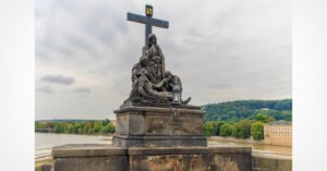 A stone sculpture of the Pietà featuring a cross stands on a pedestal on a bridge overlooking a river and a forested hillside.