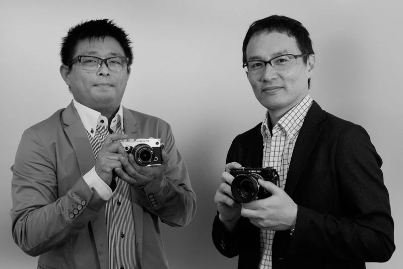 Two men in blazers and button-down shirts stand side-by-side, each holding a camera labeled "Olympus Pen" in their hands.