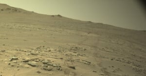 A barren, rocky Martian landscape with rover tracks stretching across the dusty, sloping terrain toward a distant ridge.