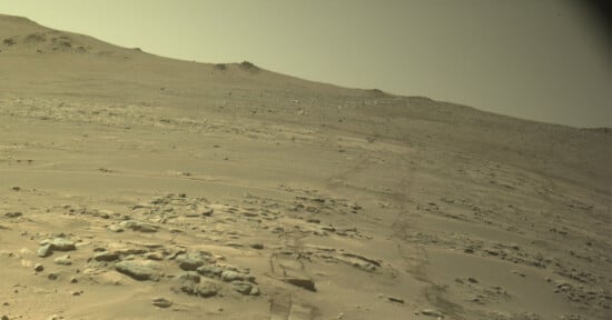 A barren, rocky Martian landscape with rover tracks stretching across the dusty, sloping terrain toward a distant ridge.