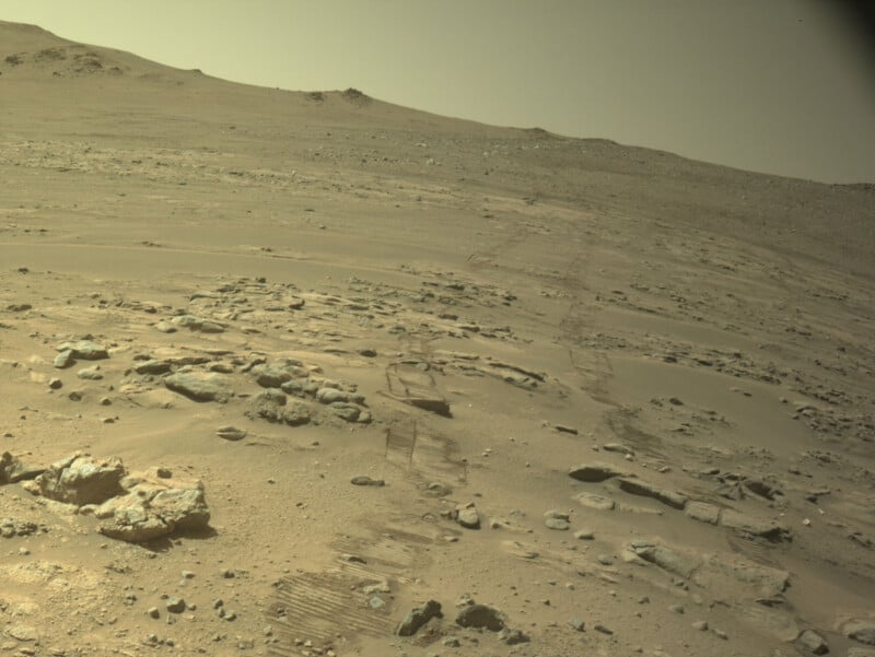 A rocky, dusty Martian landscape showing distinct parallel rover tracks stretching across the terrain toward a distant hill.