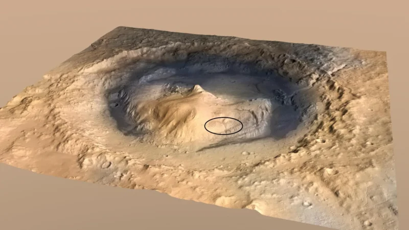 A 3D perspective view of a Martian crater featuring a central peak, with a small oval shape outlined on the slope of the mound.