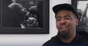 A man with a goatee and a black baseball cap smiles while speaking, positioned in front of two framed black-and-white photographs.