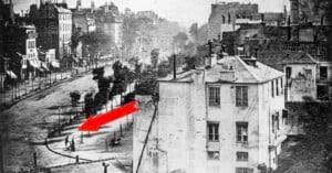 A black-and-white photograph of a Parisian street scene with a red arrow pointing toward a person having their shoes shined.