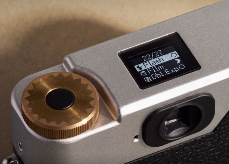 A close-up of a silver camera top plate featuring a brass shutter button and a small digital display showing menu options including Flash, Film, and Dbl. Expo.