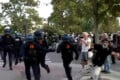 Several police officers in riot gear run toward a person in a black shirt on a paved street near trees.