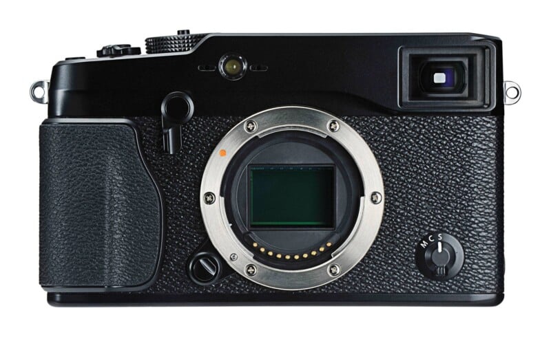 A front view of a black mirrorless digital camera body with an exposed sensor and a focus mode selector dial labeled M, C, and S.