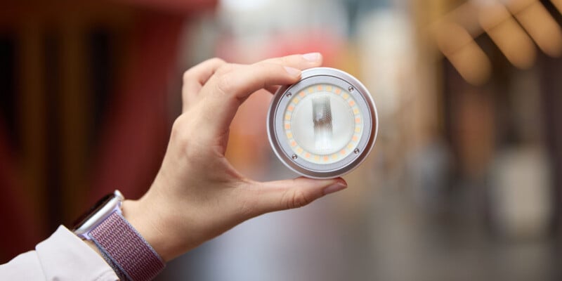 A hand holds a circular LED light fixture with a central lens and an outer ring of small light-emitting diodes.