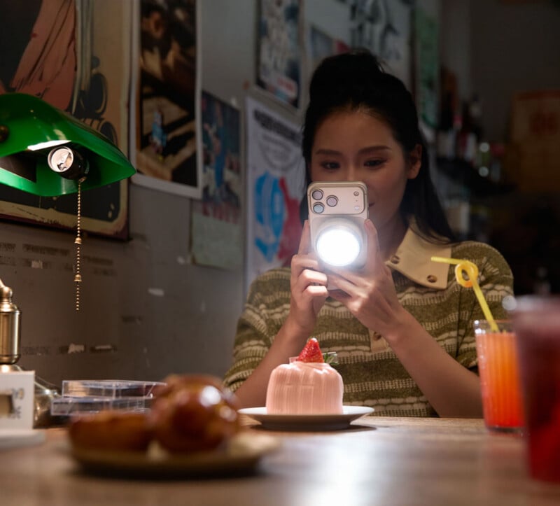 A woman in a green striped sweater uses a phone with an attached circular light to photograph a small pink cake on a table.