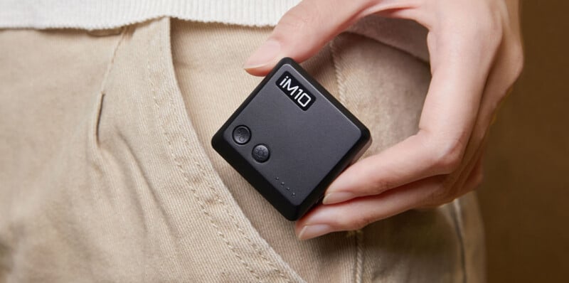 A person's hand holds a small, square, black device labeled "iM10" next to the pocket of a pair of tan pants.