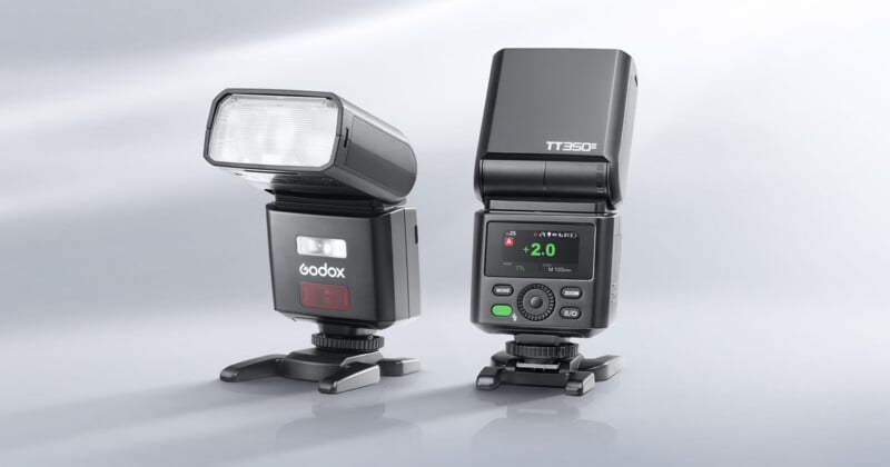 Two Godox TT350II camera flashes stand on a light gray surface, one angled to the side and the other facing forward with its digital display illuminated.