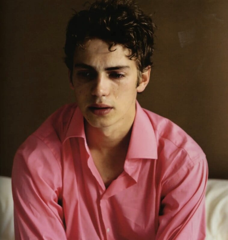 A young man with curly brown hair and a tear on his cheek wears a pink button-down shirt while looking downward.