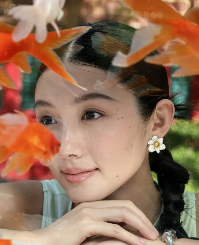 A young woman with dark hair in a braid rests her chin on her hand, framed by several goldfish swimming in the foreground.