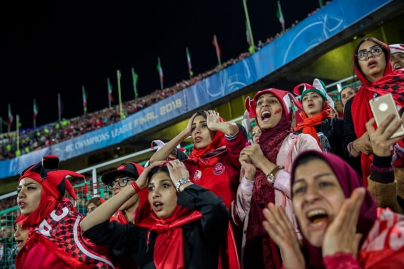 A group of women in red scarves and team gear react with excitement and tension while watching a soccer match in a stadium.