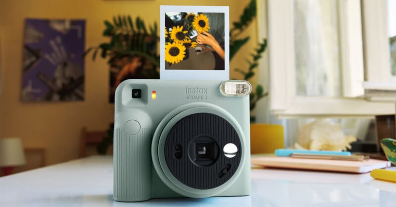 A light green instant camera sits on a white table with a developed square photo of sunflowers resting on top of it.