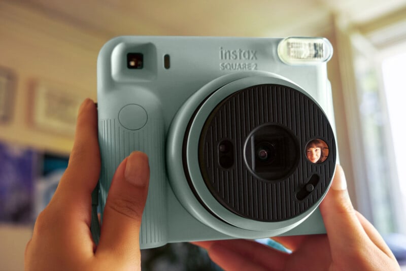 A person holds a light blue Instax Square 2 instant camera, which features a small circular mirror on the front lens housing showing a reflection of a woman's face.