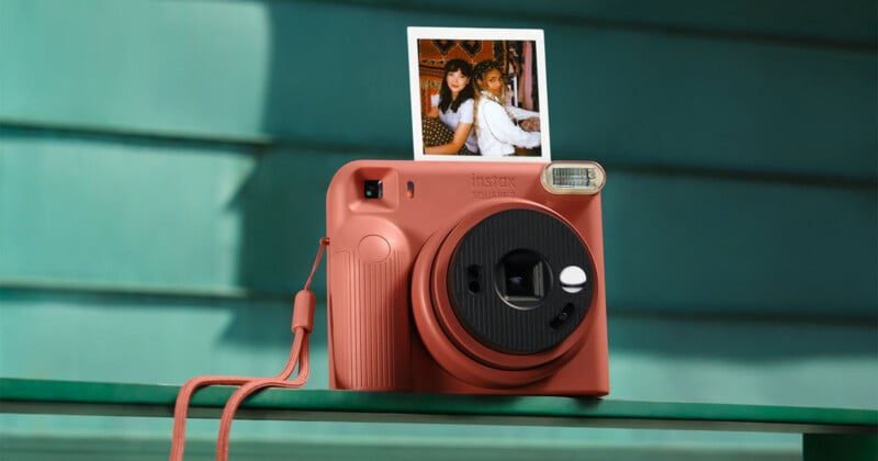 A terracotta-colored Instax Square 2 instant camera sits on a green ledge, displaying a square photograph of two people above its lens.