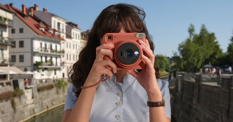 A person with long brown hair holds a terracotta-colored Instax Square 2 camera up to their face, wearing a light blue button-down shirt and a brown leather watch, with a canal and buildings in the background.