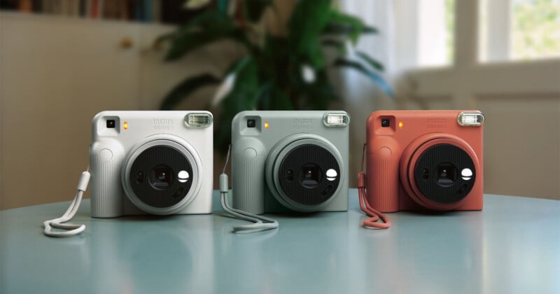 Three Instax Square 2 instant cameras in white, sage green, and terracotta orange sit side-by-side on a light blue table.