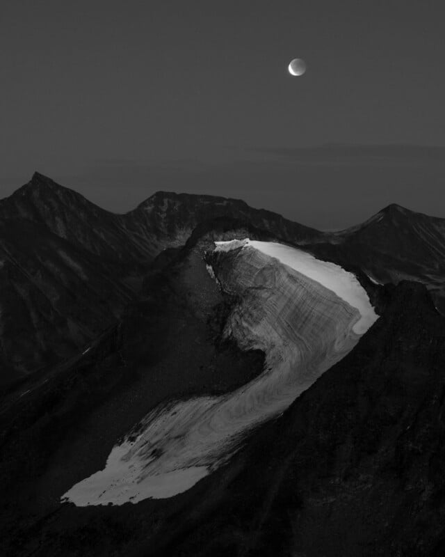 A crescent moon hangs in a dark sky above a jagged mountain range featuring a prominent, snow-covered glacier.