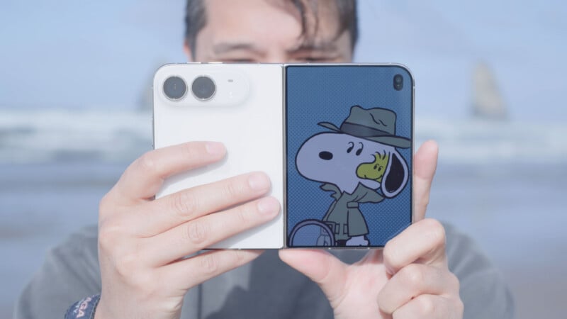 A person holds a white foldable smartphone open, displaying a cartoon of Snoopy as a detective on the screen, with a blurry beach scene in the background.