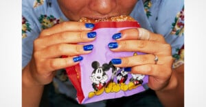 A person with blue glittery nail polish holds a burger in a purple Mickey Mouse wrapper while taking a bite.