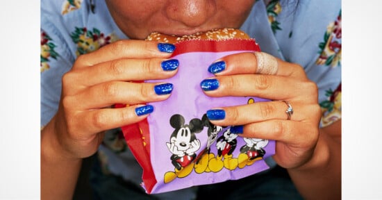 A person with blue glittery nail polish holds a burger in a purple Mickey Mouse wrapper while taking a bite.