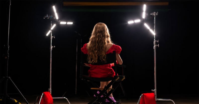 A young girl with long wavy hair, wearing a red dress, sits on a director's chair between two bright studio lights against a black background.