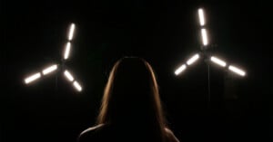 A person with long hair is silhouetted against a dark background between two tripod-mounted, three-armed LED light fixtures.