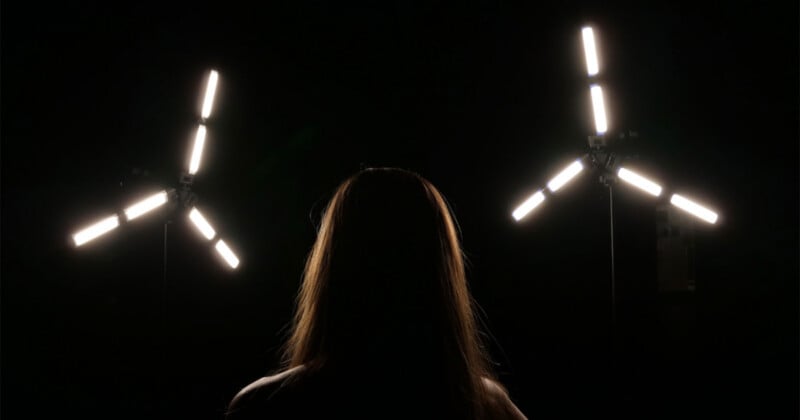 A person with long hair is silhouetted against a dark background between two tripod-mounted, three-armed LED light fixtures.