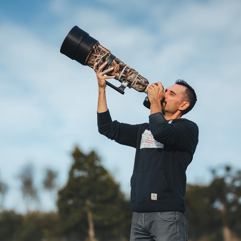 A man in a dark hoodie holds a large telephoto lens with a camouflage cover, aiming it upward toward the sky.