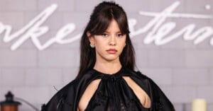 Mia Goth with dark hair and bangs wears a black satin dress with cutouts and ornate earrings in front of a brick wall.