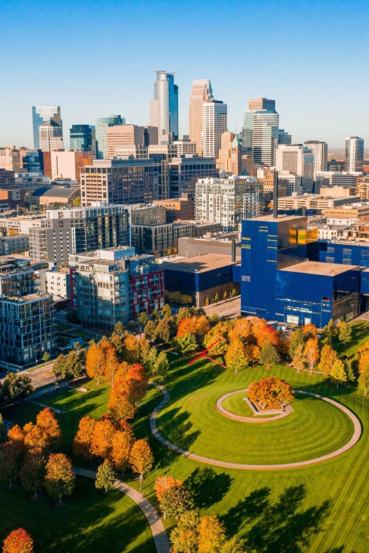 An aerial view of a city skyline overlooking a park with autumn trees, a spiral walking path, and a large blue building.