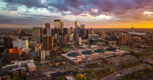 An aerial view of a city skyline at sunset, featuring numerous skyscrapers, a large convention center with domed roofs, and a highway in the foreground.