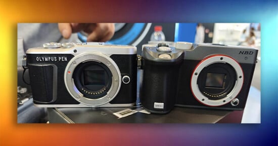 Two digital cameras sit side-by-side on a surface, with the left one labeled "OLYMPUS PEN" and the right one labeled "NBD" and "G500".
