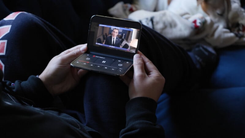 A person holds a foldable smartphone in a half-open position, displaying a video playback screen with controls on the bottom half.