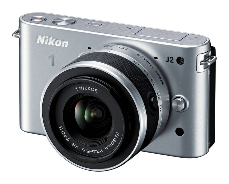 A silver Nikon 1 J2 mirrorless camera with a 10-30mm lens attached, viewed from the front-right angle against a white background.