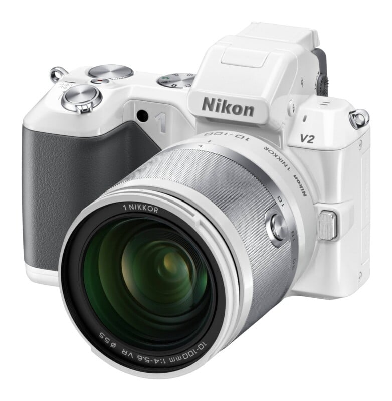 A white Nikon V2 mirrorless camera with a silver 10-100mm lens attached, set against a plain white background.
