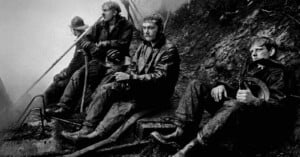 Four firefighters in dark, soot-covered gear sit exhausted on a rocky, smoky hillside, resting with their helmets in hand.