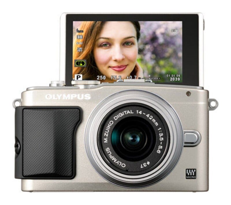 A silver Olympus mirrorless camera with a 14-42mm lens and a flip-up screen displaying a portrait of a woman.