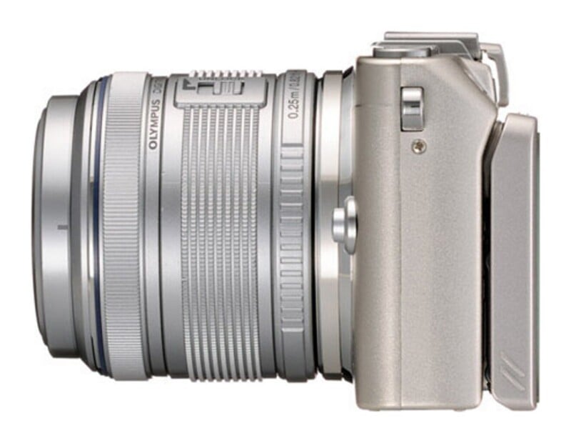 A side profile of a silver mirrorless digital camera with an attached lens, showing the lens barrel, focus ring, and camera body.