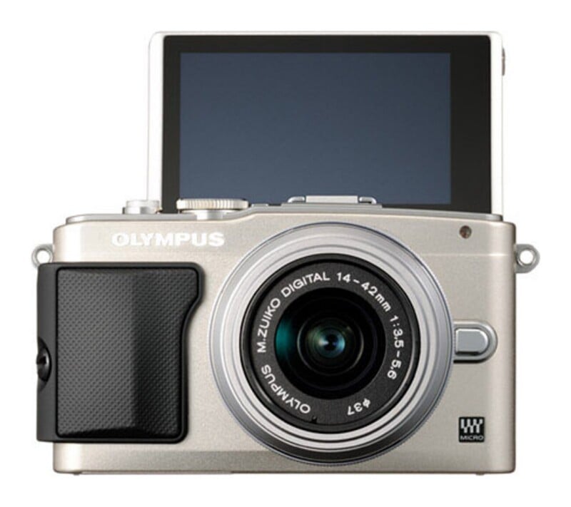 A silver Olympus mirrorless camera with a lens attached and its rear screen flipped upward.