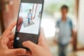 A person holds a smartphone to take a photo of a young boy standing in a doorway, while the boy is also visible in the background, out of focus.
