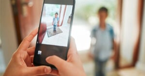 A person holds a smartphone to take a photo of a young boy standing in a doorway, while the boy is also visible in the background, out of focus.