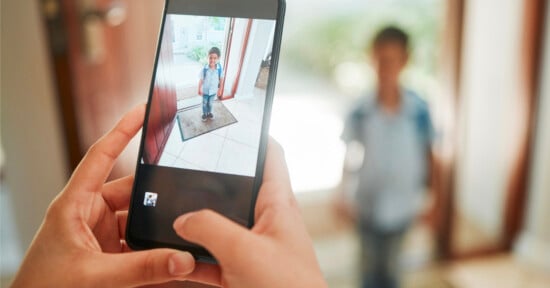 A person holds a smartphone to take a photo of a young boy standing in a doorway, while the boy is also visible in the background, out of focus.