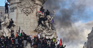 A large group of protesters climbs and stands on the base of a stone monument in Paris, with thick smoke rising in the background.