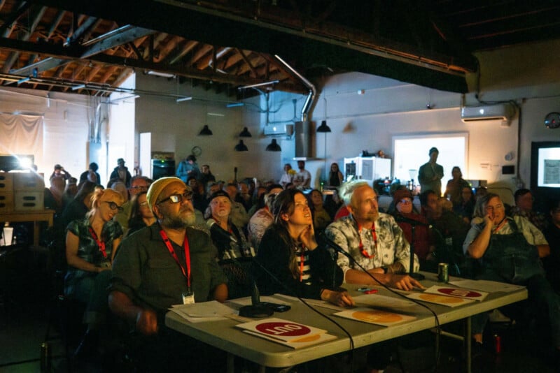 A panel of three people sits at a table facing an audience in a dimly lit, industrial-style room with exposed wooden rafters.